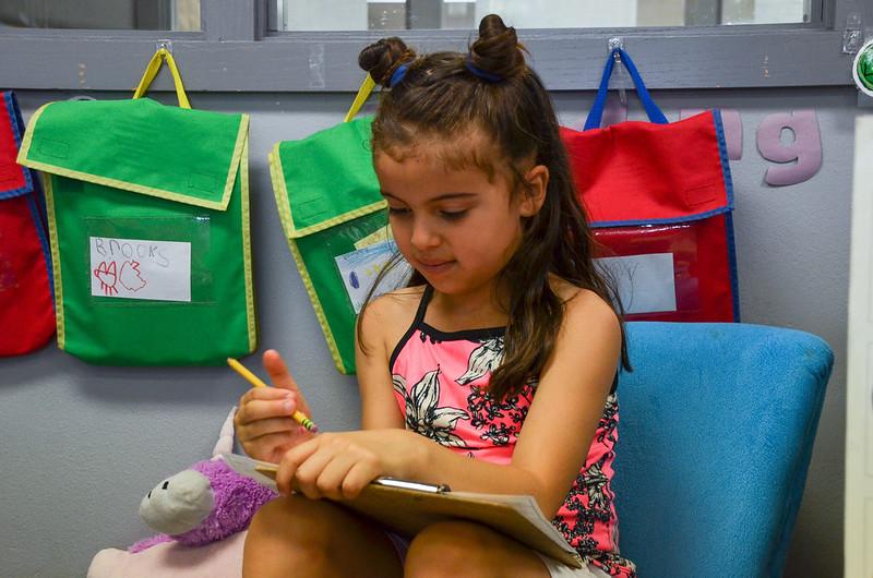 A child sits in a comfortable chair and uses a pencil and clipboard with paper to write.
