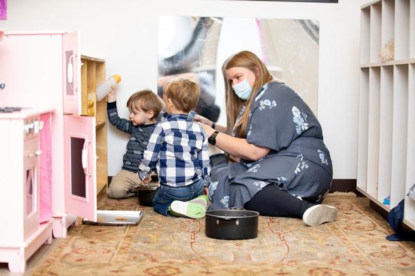 A teacher and two children are interacting while sitting on the floor in the housekeeping area.