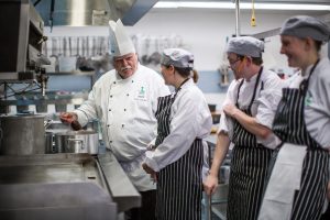 Culinary faculty member dressed in chef uniforms, gather around a stove in a kitchen, demonstrating culinary techniques to students.