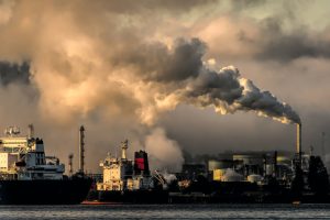 Image of some ships and then a factory in the background with large amount of smoke going into the sky.