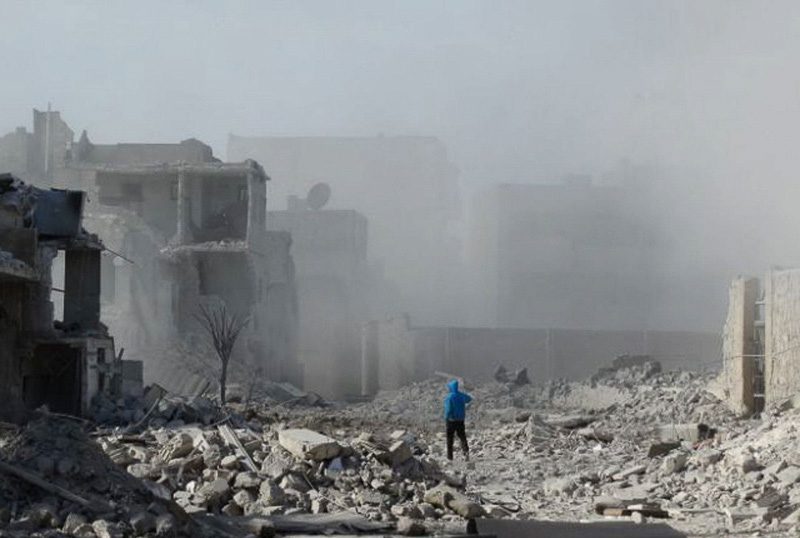 Person in blue hooded jacket standing in the rubble of a destroyed building.