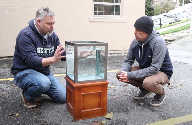 Two people, Mark and Chad, crouched down talking with a container filled with one cubic foot of water sitting between them.