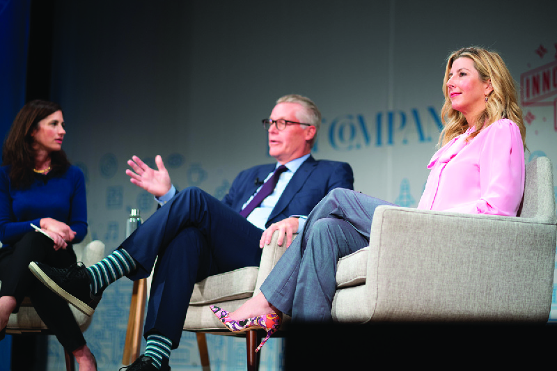 Two women and a man are seated on a couch at a panel presentation. The man is speaking and gesturing with one hand.