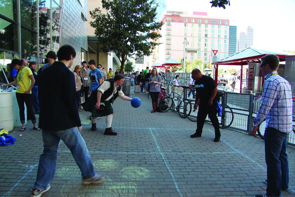 Four adults are outside on a brick sidewalk playing Foursquare with a blue ball while others look on.