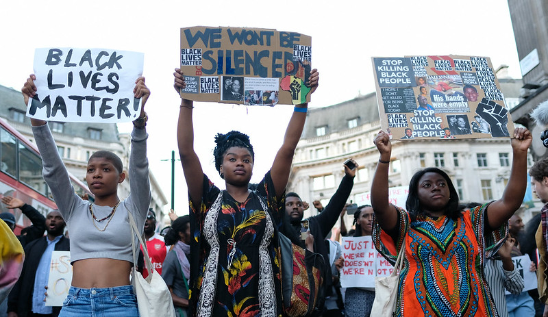 Three Black women holding signs at a demonstration: "Black Lives Matter," "We won't be silenced," "Stop killing Black people."