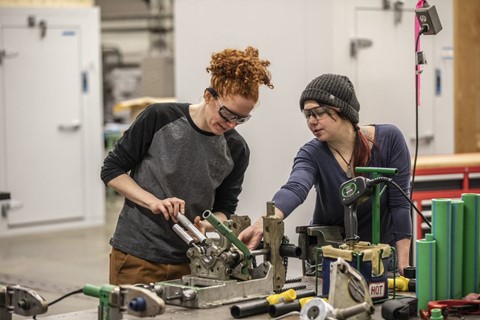 One woman works at a workbench while the offer points to offer suggestions.