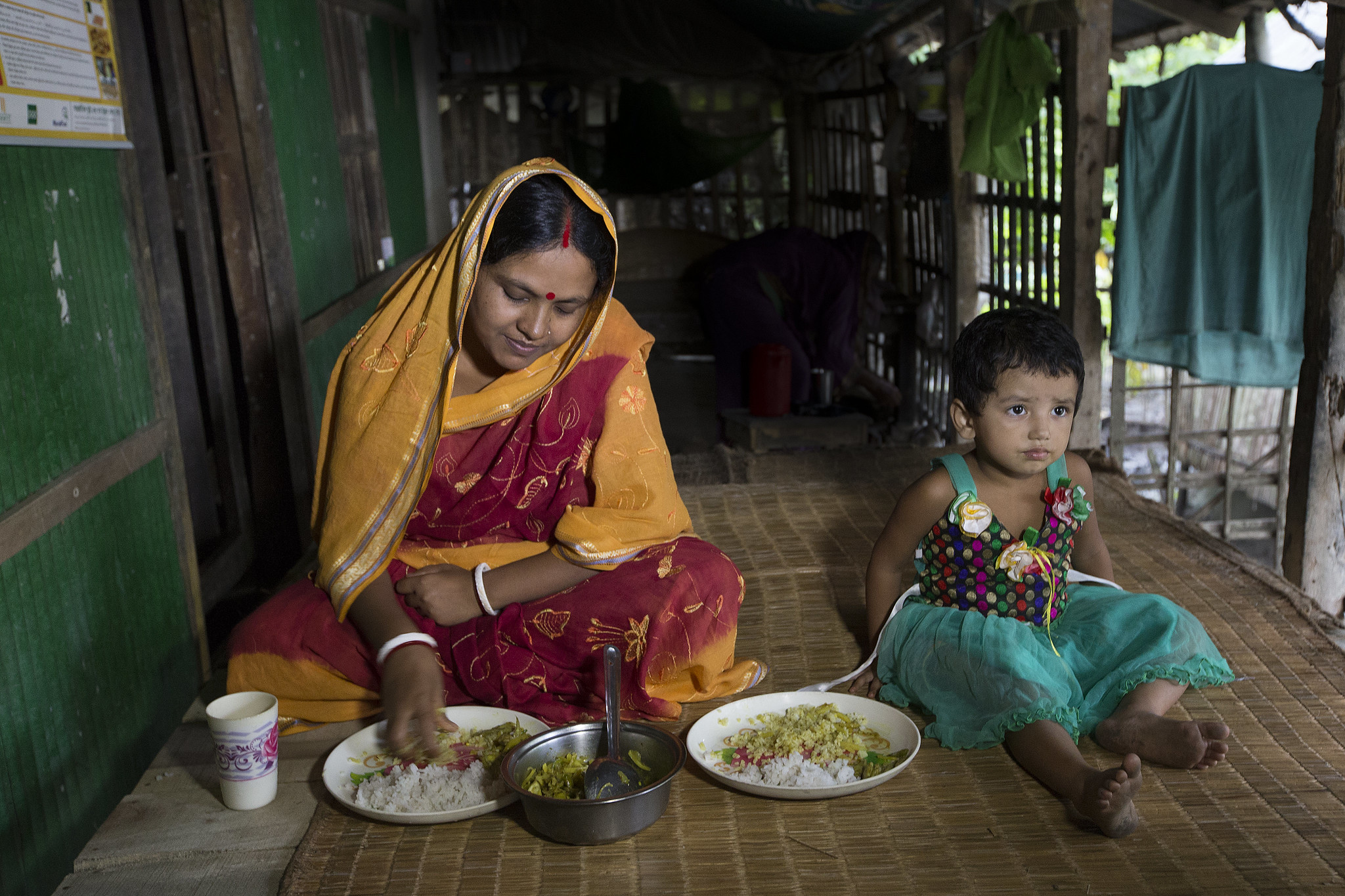 Mother and her daughter in Khulna Bangladesh, sit cross-legged on the floor eating a rice-based dish.