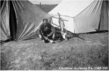 Black and white photograph showing Maslow sitting cross-legged on the ground in front of traditional housing
