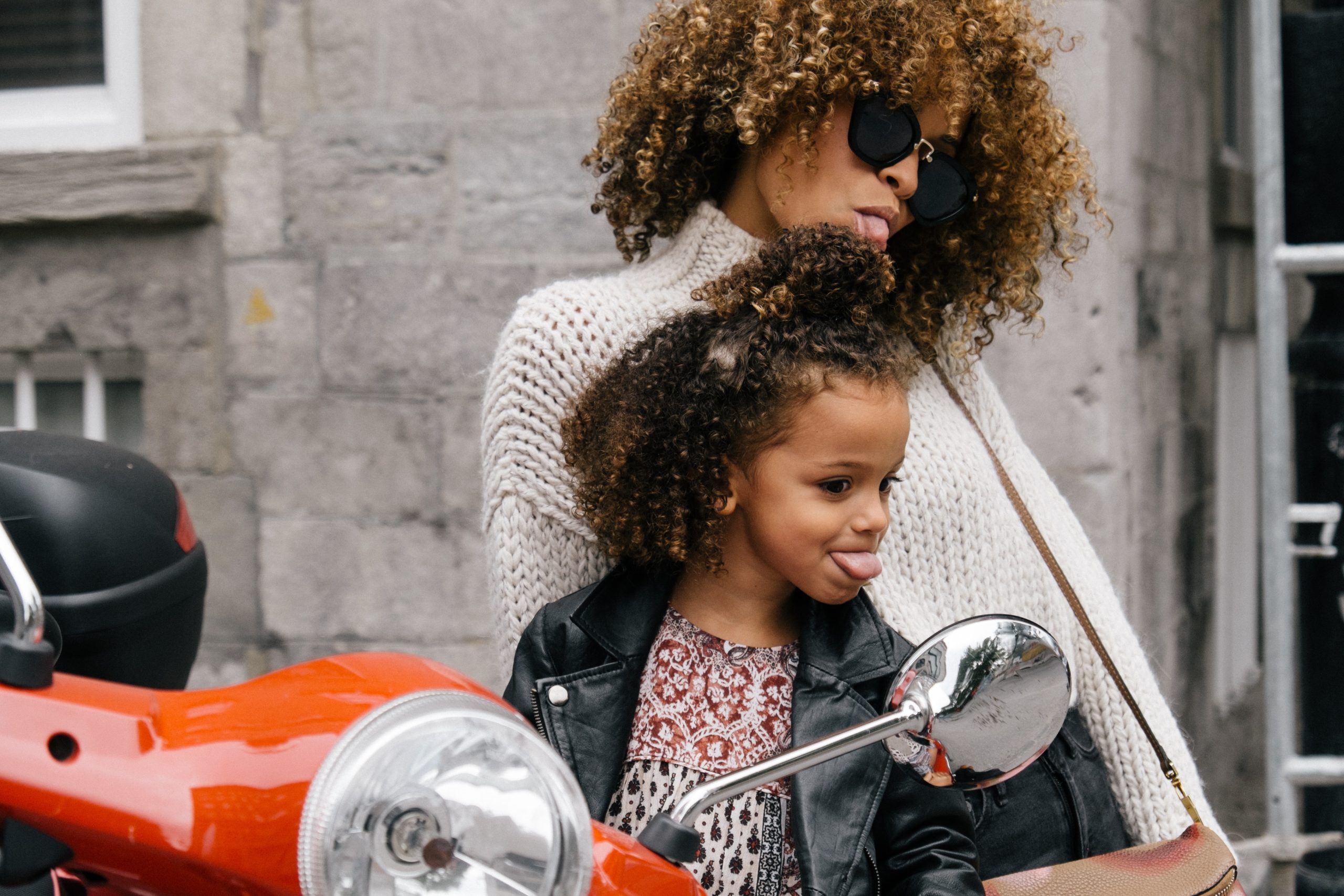 A parent sits on a motorcycle with their young child, both look into the rearview mirror, sticking out their tongues.