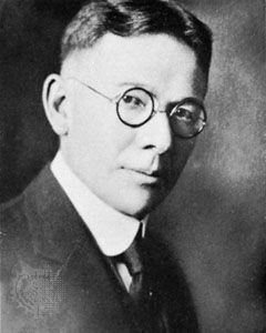Public domain, via Wikimedia Commons Black-and-white portrait of a man wearing round glasses, a suit, and a tie, looking slightly to the side with a serious expression.