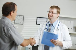 A healthcare professional, wearing a white coat and stethoscope, greets a patient with a handshake in a clinical office. The doctor holds a clipboard and smiles warmly, suggesting a positive rapport and the start of a therapeutic or assessment relationship. The setting includes typical office elements such as binders, a clock, and framed certificates, reinforcing the professional context