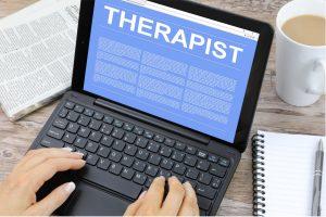 Therapist by Nick Youngson CC BY-SA 3.0 Pix4free.org Person typing on a laptop with the word 'THERAPIST' displayed in large white letters on the screen. The laptop sits on a wooden desk beside a folded newspaper, a white mug of coffee, and an open spiral notebook with a pen.
