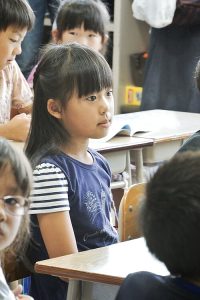 MIKI Yoshihito from Sapporo City, Hokkaido, Japan, CC BY 2.0 <https://creativecommons.org/licenses/by/2.0>, via Wikimedia Commons A young girl sits attentively at her desk in a classroom, surrounded by other children. She is facing forward and appears focused, possibly listening to the teacher or engaging in a classroom activity. Open notebooks and books are visible on the desks, indicating an academic setting. The image captures a moment suitable for behavioural observation in an educational environment.