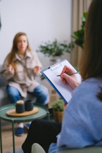Courtesy of Hippopx (Free for commercial use) A therapist is sitting across from a client during a counseling session in a calm, well-lit room. The therapist, partially visible in the foreground, is holding a clipboard and writing notes. The client, seated across from them, appears to be speaking and gesturing with her hands. A small round table with two mugs and a teapot sits between them, and the background includes plants and soft furnishings, contributing to a comfortable, private setting.