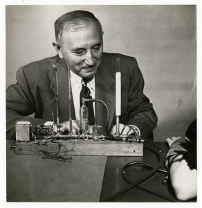 Black-and-white photograph of an older man in a suit, smiling while seated at a table with a scientific or experimental apparatus. The device includes glass tubes, wires, and a vertical gauge. A person’s arm with an attached cuff and tubes is visible on the right side, suggesting a physiological measurement experiment.