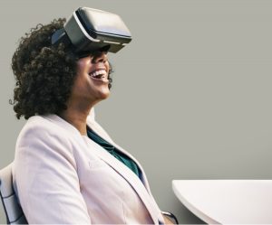 Courtesy of pxhere Smiling woman wearing a virtual reality headset while sitting indoors. She appears to be enjoying the VR experience, dressed in a light blazer and dark shirt.