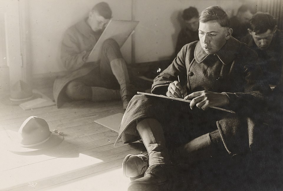 Black-and-white photo of young men in military uniforms sitting on the floor and writing on large sheets of paper. One man in the foreground looks focused as he writes with a pencil on a clipboard resting on his knee. This image depicts soldiers taking the Army Alpha Test during World War I.