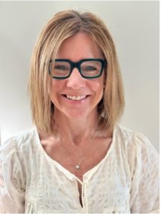 Smiling woman with shoulder-length light brown hair wearing rectangular dark-framed glasses and a white blouse. She is also wearing a delicate necklace with a small pendant.