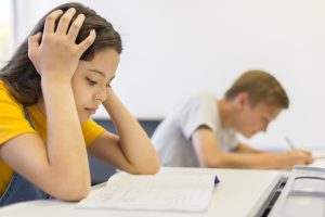 Student in a yellow shirt sits at a desk with hands on head, looking stressed over an open notebook; another student in the background writes, out of focus.