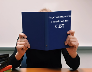 Image adapted by Madison Conrad from a free-to-use image on PickPik (no author listed, public domain). Person sitting at a desk holding a blue book that covers their face. The book’s cover text reads: ‘Psychoeducation: a roadmap for CBT.’ Several other books are stacked on the desk to the side.