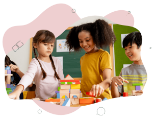 Three children standing and laughing at a desk in the classroom building structures with blocks.