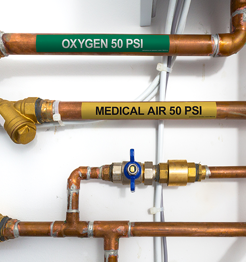 Several copper gas pipes, two of which with labels on them. One label is green and reads “oxygen 50 PSI”. The other label is yellow and reads “medical air 50 PSI”.